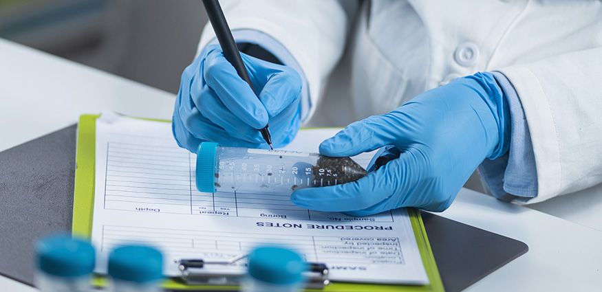 Scientist wearing blue latex gloves and writing on a vial of cannabis products for lab testing. 